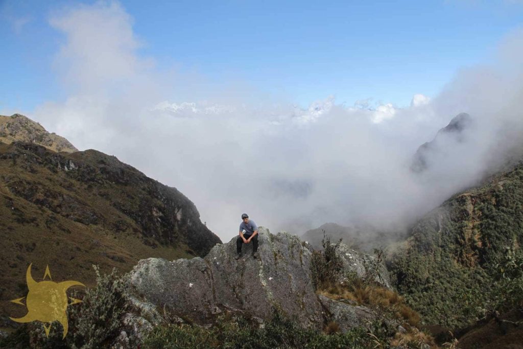 A trekker on a rock outcrop above a sea of cloud, with snow peaks on the horizon