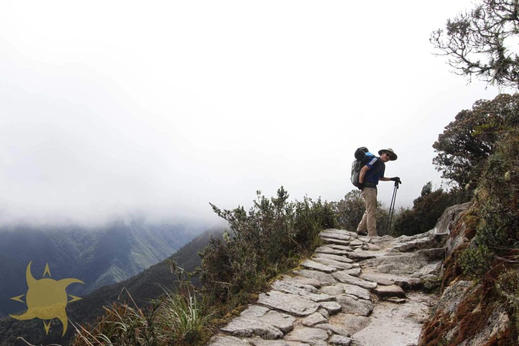 A trekker with poles and a full pack pausing on the Inca stone trail along a cliff edge above cloud